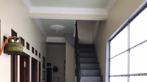 A hallway with a staircase leading to an upper floor. There are doors on the left, a window on the right, and a trash bin near the stairs. - Villa Bintang (Malang)