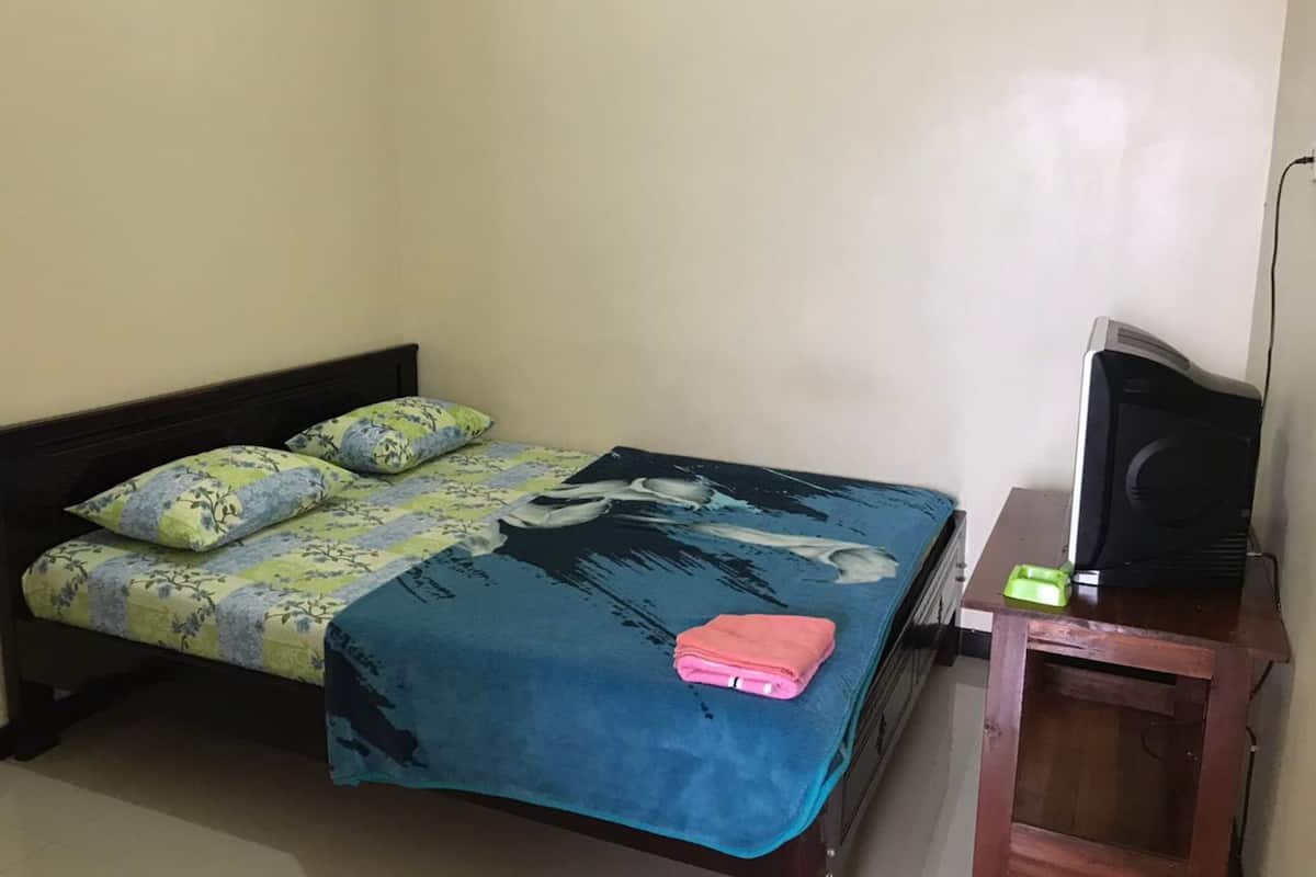 Una cama individual con ropa de cama floral, una manta azul y una toalla rosa. Un televisor está sobre un soporte de madera junto a la cama.