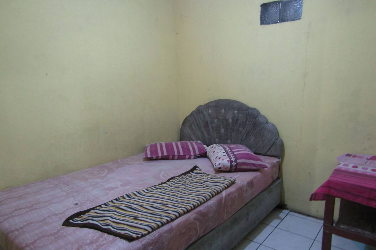 A small, single-bed room with a plain wall, a bed with a pink bedspread and striped pillow, and a small table with a pink tablecloth.