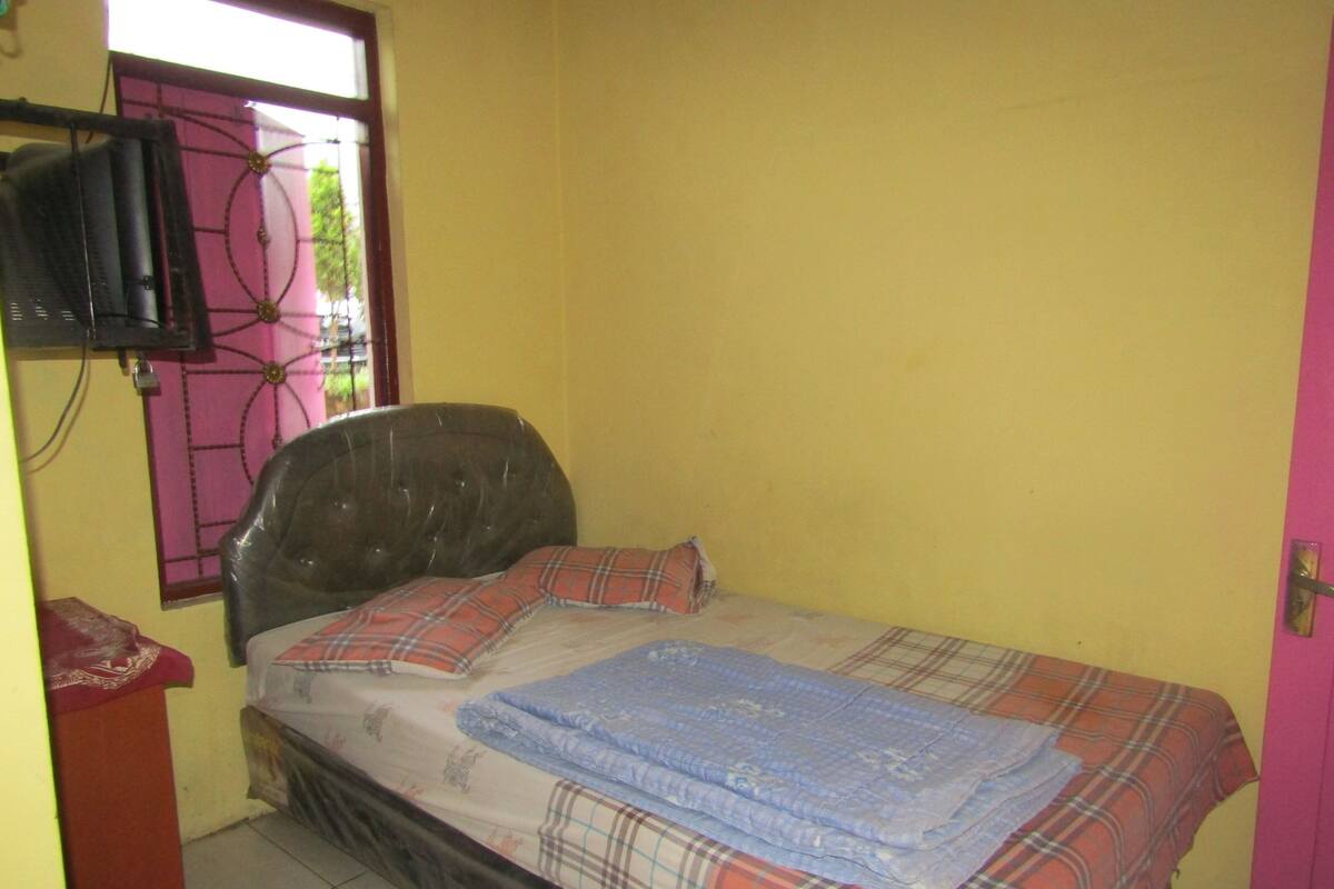 A small, single-bed room with a wooden headboard, a window with a decorative grille, and a pink door.
