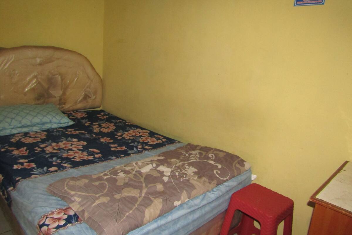 A small, single-bed room with a floral bedspread, a red plastic stool, and a wooden nightstand.