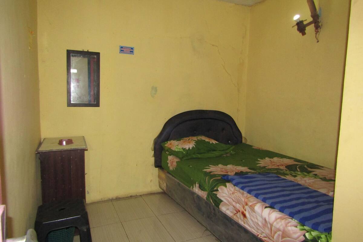 A small, yellow-walled room with a single bed, a small wooden cabinet, and a mirror on the wall.