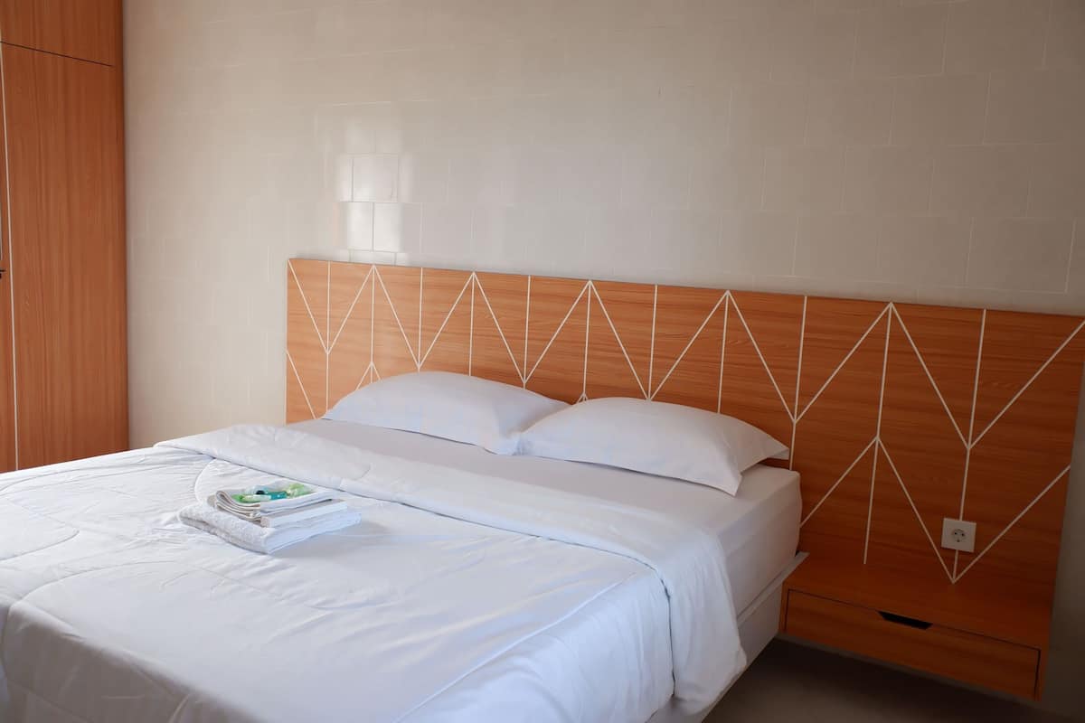 Een net opgemaakt bed met witte lakens, een houten hoofdbord met een geometrisch patroon, en een klein nachtkastje met een tijdschrift.