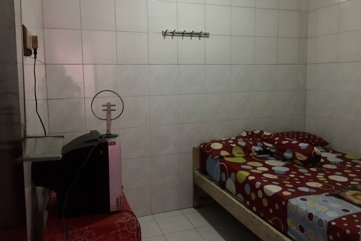Een kleine, zwak verlichte kamer met witte betegelde muren, een bed met een stippenbedekking, een bureau met een computer en een radiator aan de muur.