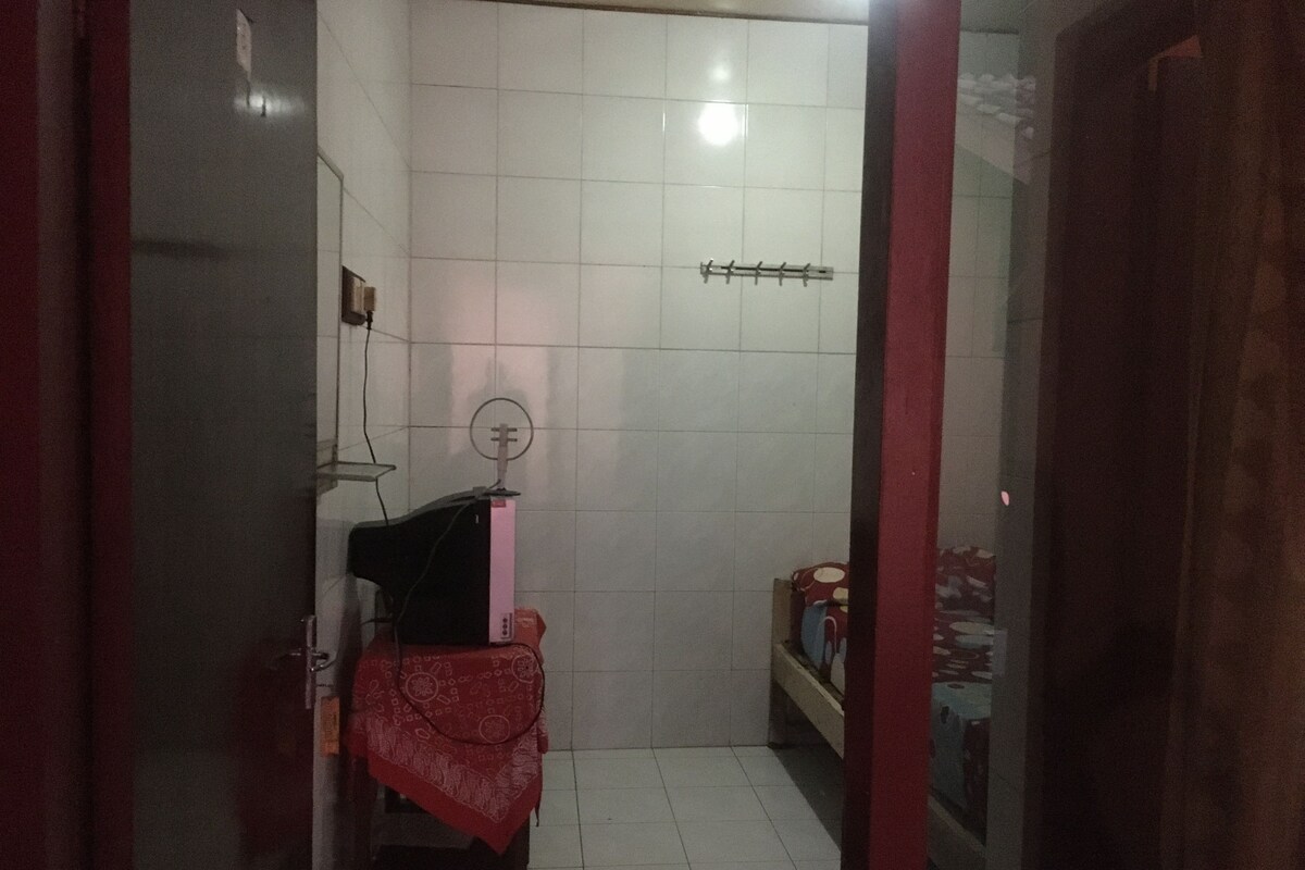 Une petite pièce faiblement éclairée, aux murs recouverts de carreaux blancs, avec une seule chaise, une table recouverte d’une nappe rouge et une petite étagère sur laquelle sont posés quelques objets.