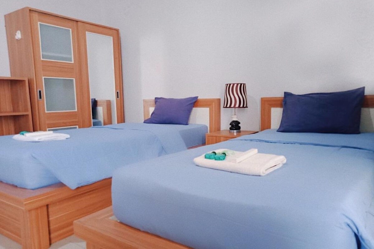 A hotel room with two beds, each with a blue bedspread and pillows, a wooden wardrobe, and a nightstand with a lamp.