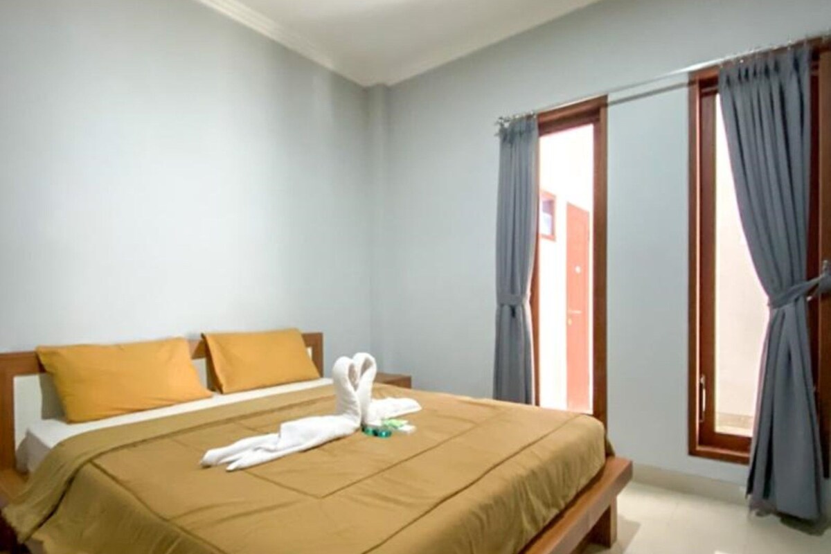 A hotel room with a large bed, two pillows, a wooden headboard, a window with curtains, and a towel arrangement on the bed.