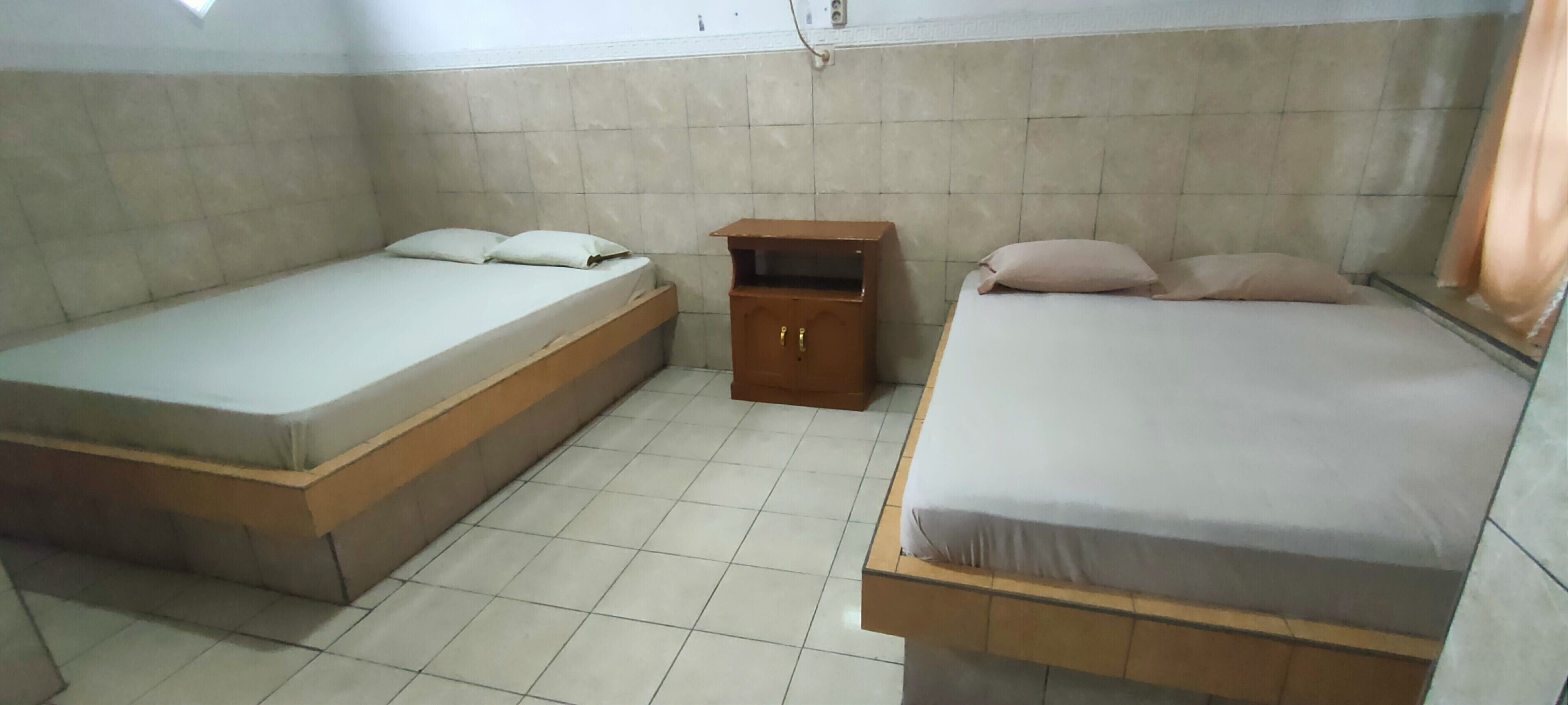 Una habitación pequeña con dos camas, una mesita de noche de madera y suelo de baldosas.