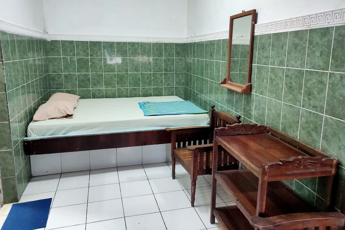 Una habitación pequeña con cama individual, paredes de azulejos verdes, un banco de madera y un espejo.
