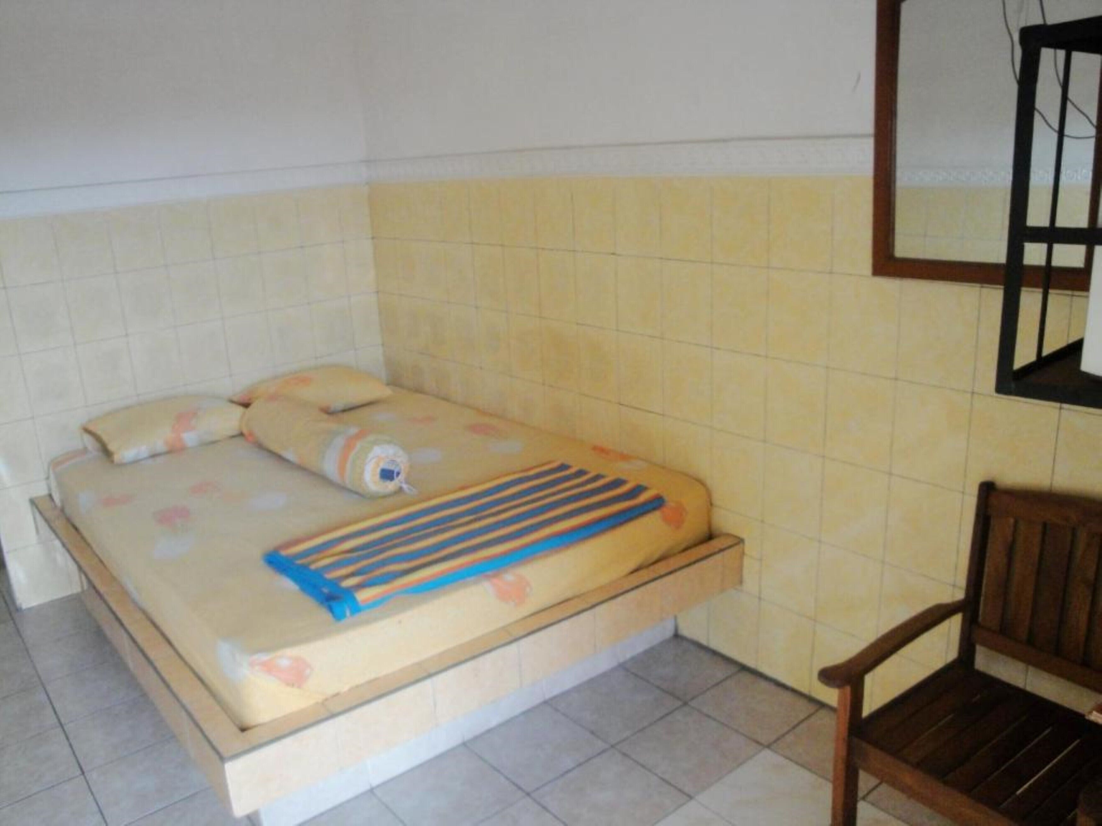 Una habitación pequeña con cama individual, una silla de madera y una ventana con marco de metal.