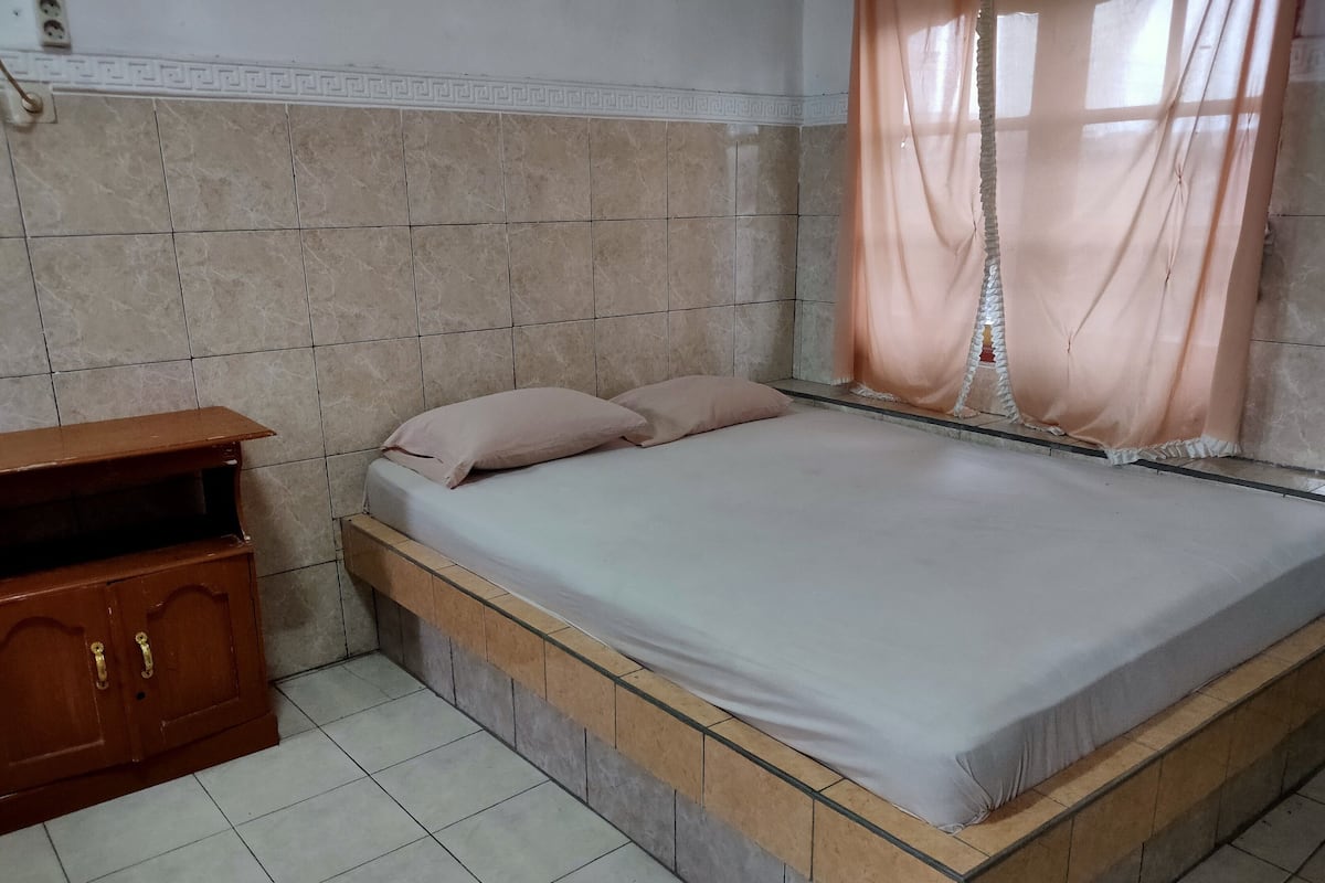 Un dormitorio con cama, un aparador de madera y una ventana con cortinas traslúcidas.