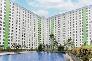 A high-rise residential building with multiple balconies, a swimming pool, and palm trees. - Juragan Room (South Tangerang)