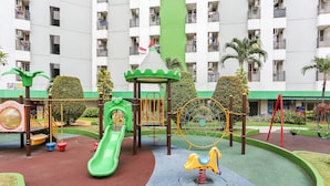 A playground with a slide, swings, and a climbing structure, surrounded by a green border and palm trees. - Juragan Room (South Tangerang)