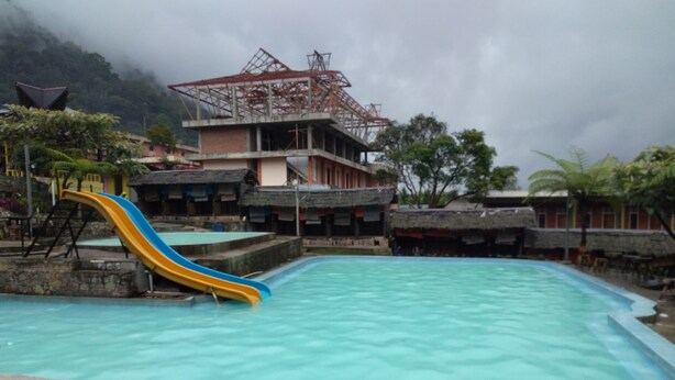 A swimming pool with a blue and yellow slide, surrounded by buildings and trees. - Penginapan Puncak DP (Berastagi)