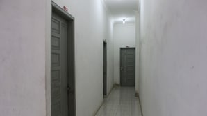 A narrow hallway with white walls and tiled flooring. There are two closed doors on either side, one with a red sign above it. - Penginapan Transit Tomato (Medan)