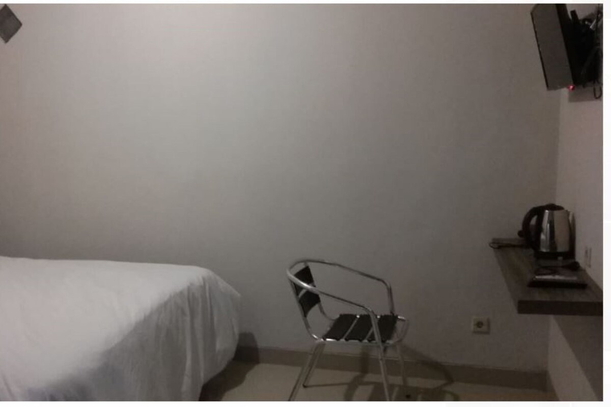 A small room with a bed, a chair, and a shelf with a kettle.