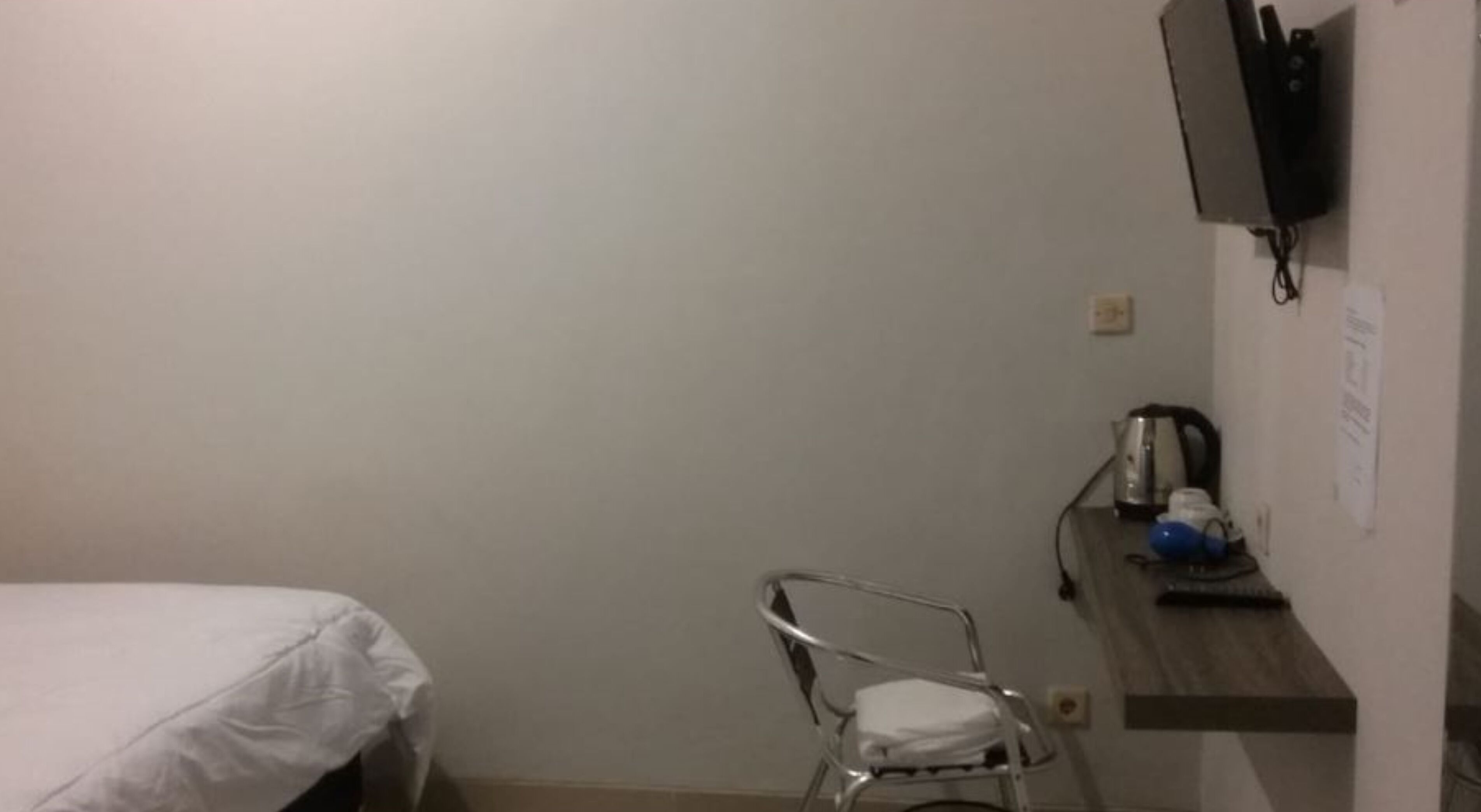 A small, sparsely furnished room with a bed, a chair, a TV mounted on the wall, and a small shelf with a kettle and other items.