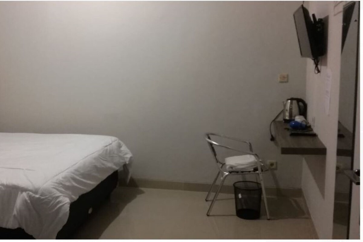 A small, simple room with a bed, a chair, a trash bin, and a small table with a kettle and a cup.