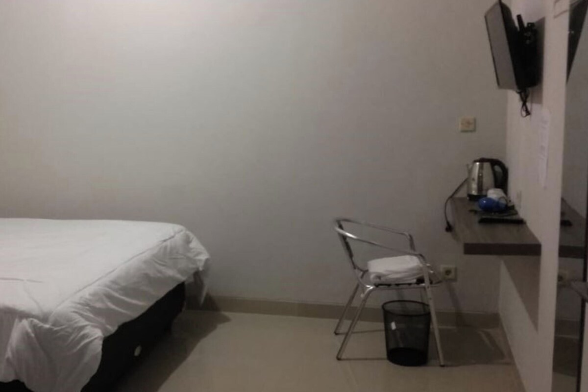 A small, sparsely furnished room with a single bed, a chair, a small table, and a wall-mounted TV.