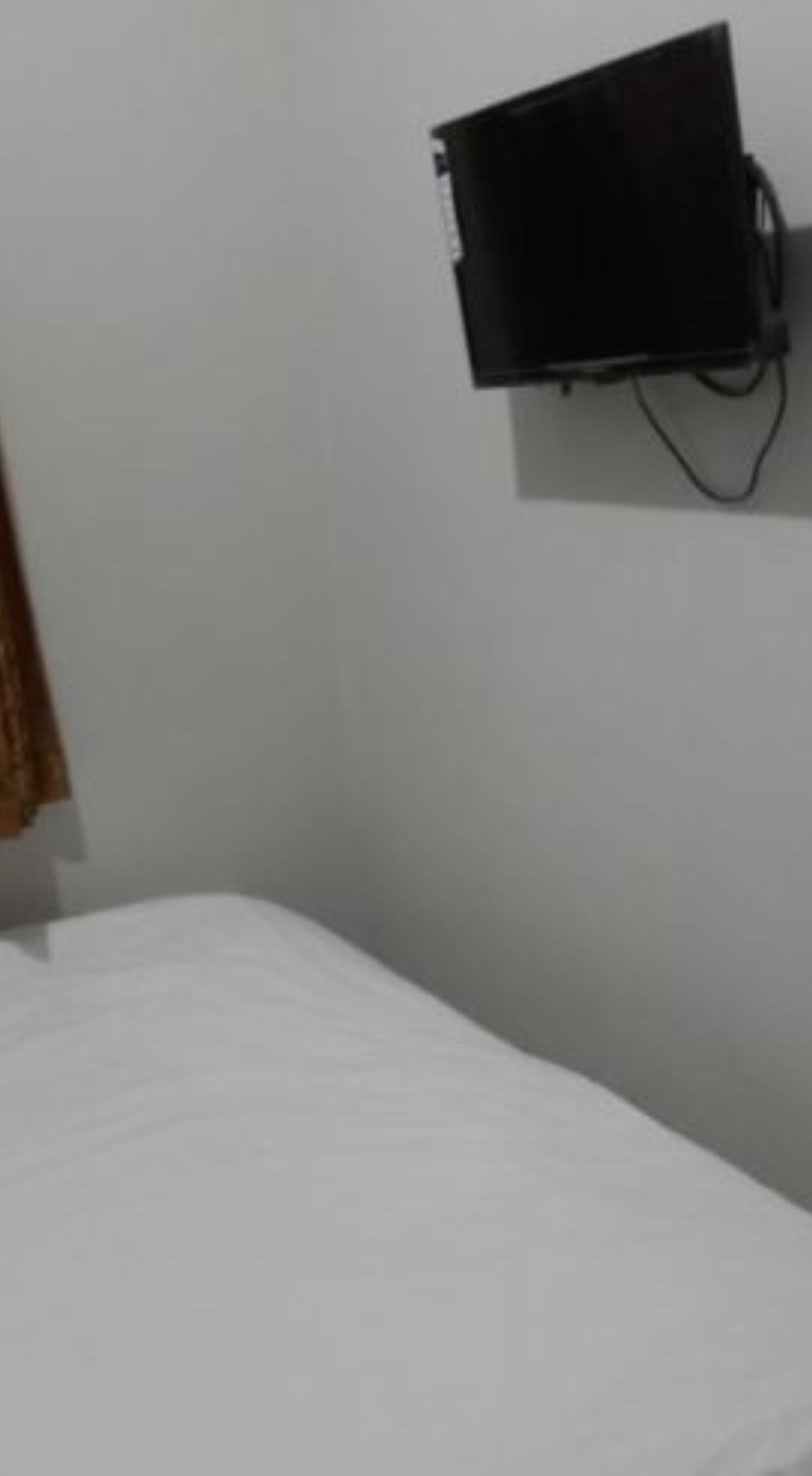A room with a white bed, a wall-mounted TV, and a curtain on the left.