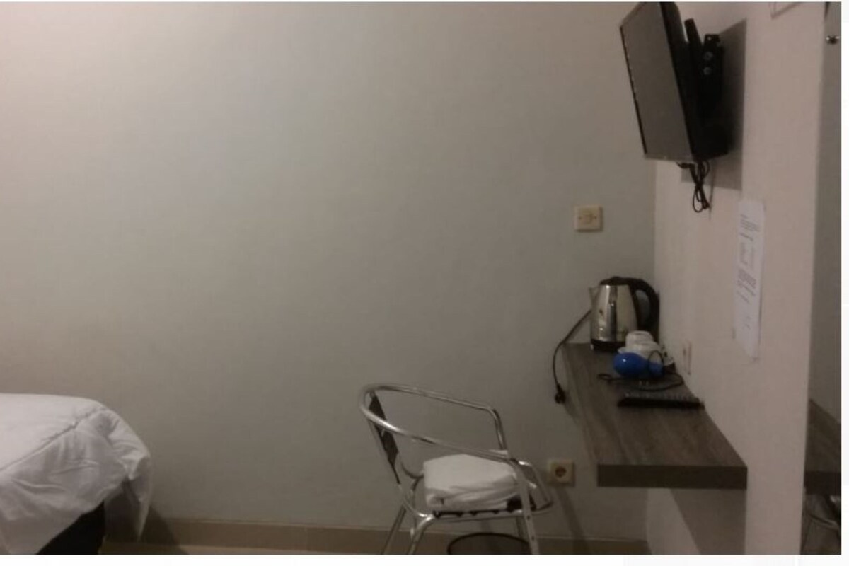Ein schlichter, einfach eingerichteter Raum mit einem Bett, einem Stuhl, einem Schreibtisch mit Wasserkocher und einem an der Wand befestigten Fernseher.