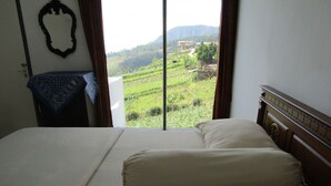 A bedroom with a large bed, a view of a green landscape through a window, and a wooden headboard. - Hotel Pantes (Plaosan)