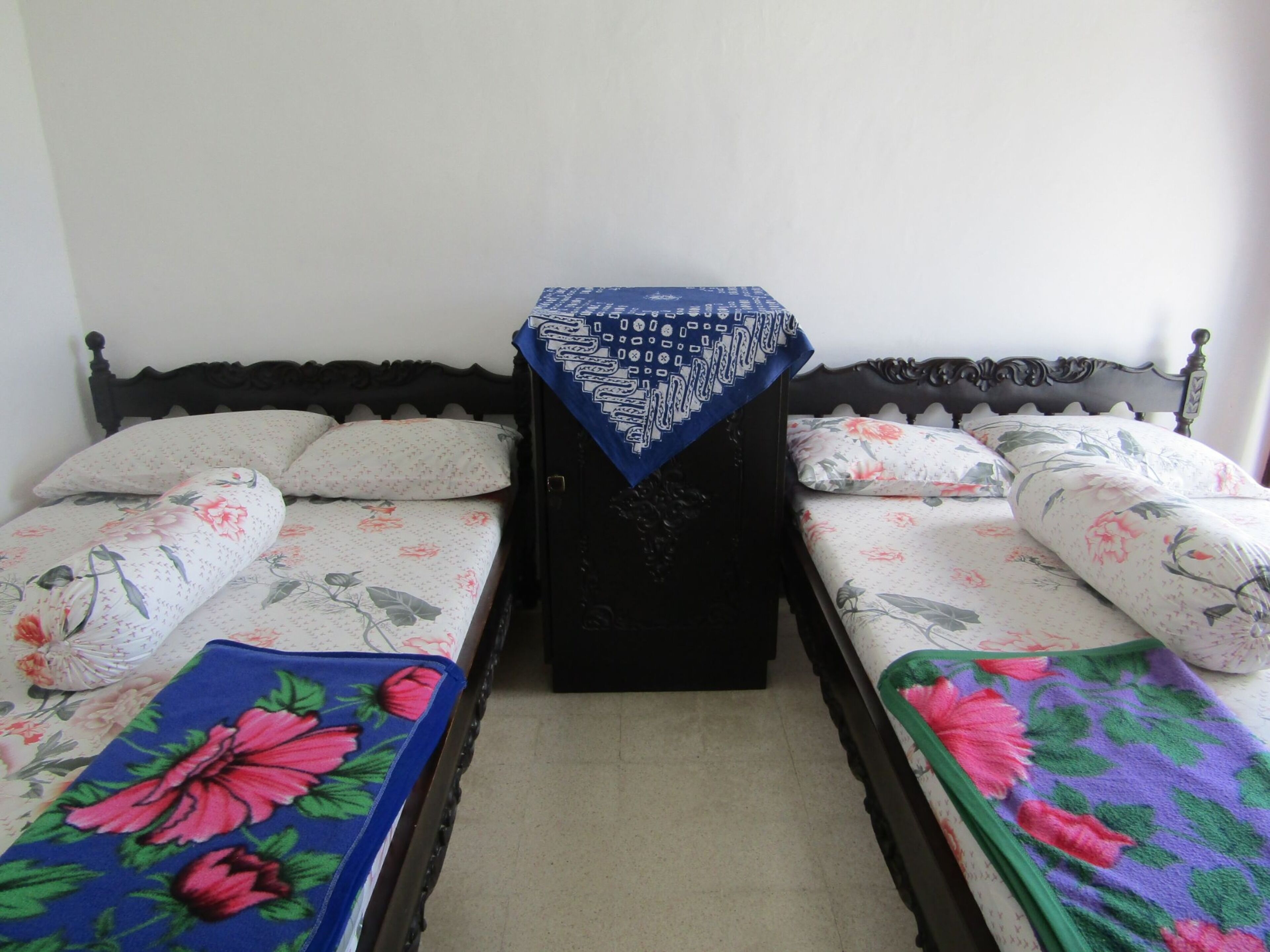 A room with two beds, each with floral bedding and a matching pillow. There is a small table with a blue cloth in the center.
