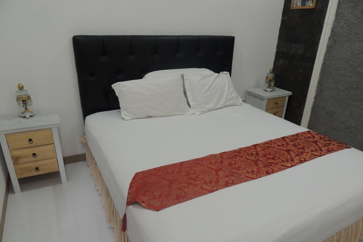 A neatly made bed with white linens and a red patterned throw, flanked by two bedside tables with lamps and a small drawer.