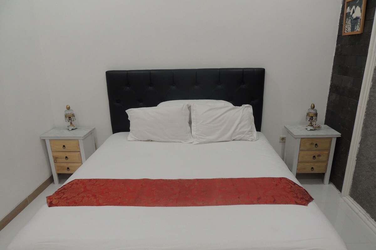 A bed with a black headboard, white pillows, and a red blanket. There are two bedside tables with lamps and drawers.
