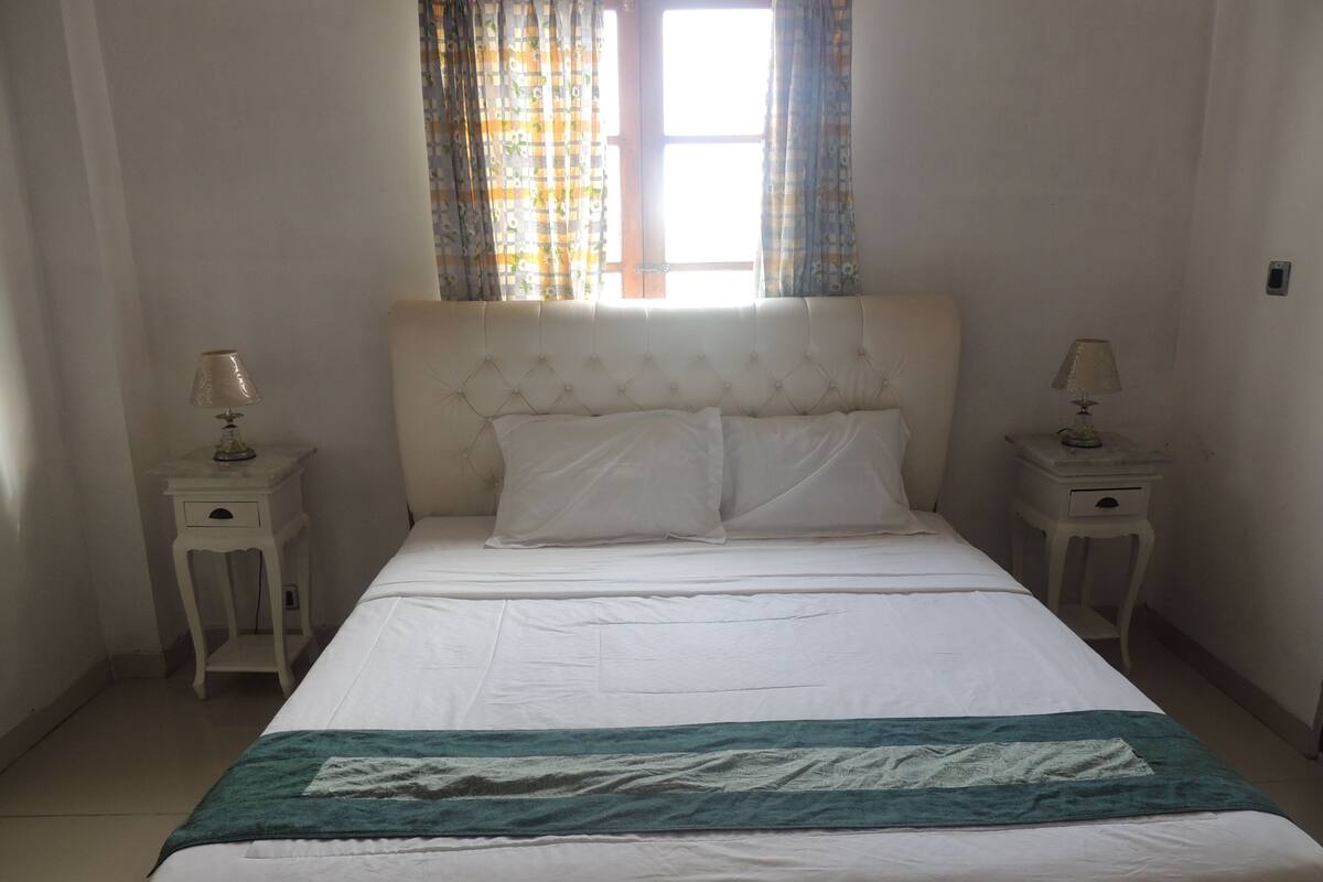 A neatly made bed with white linens and a teal blanket, flanked by two bedside tables with lamps, in a room with plain walls and a window with patterned curtains.