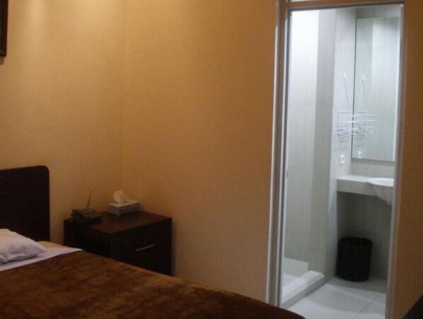 Habitación de hotel con una cama, una mesita de noche con un box de pañuelos, un cajón pequeño y una puerta de cristal que da a un baño con un lavabo y un cubo de basura.