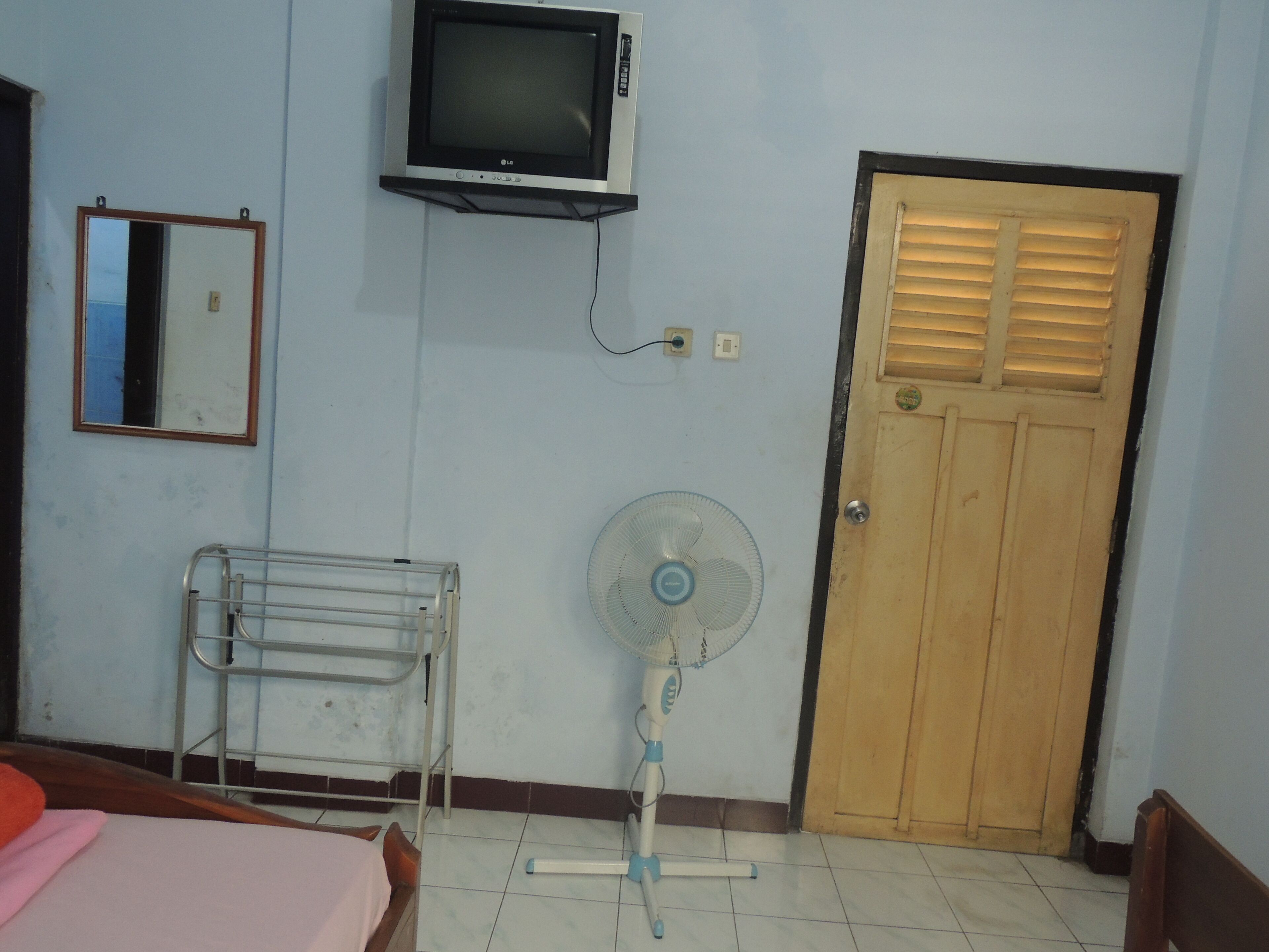 A room with a bed, a TV mounted on the wall, a wooden door, and a fan.