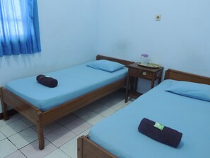A room with two single beds, each with a blue mattress and a dark-colored pillow. There is a small wooden nightstand with a lamp and a tray with a jar and a glass. - Hotel Sejahtera (Kebumen)