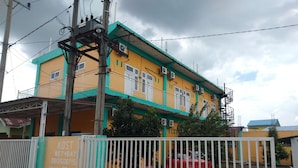 A multi-story building with a yellow and green facade, a gated entrance, and a red car parked in front. - KOST 748 AT (Medan)