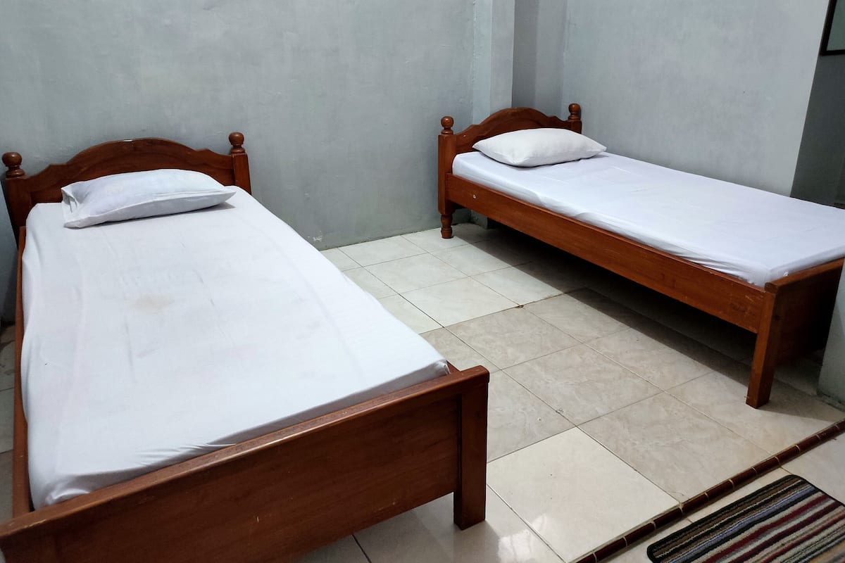 Deux lits simples en bois, recouverts de draps blancs, dans une pièce dont le sol est carrelé et les murs sont de couleur unie.