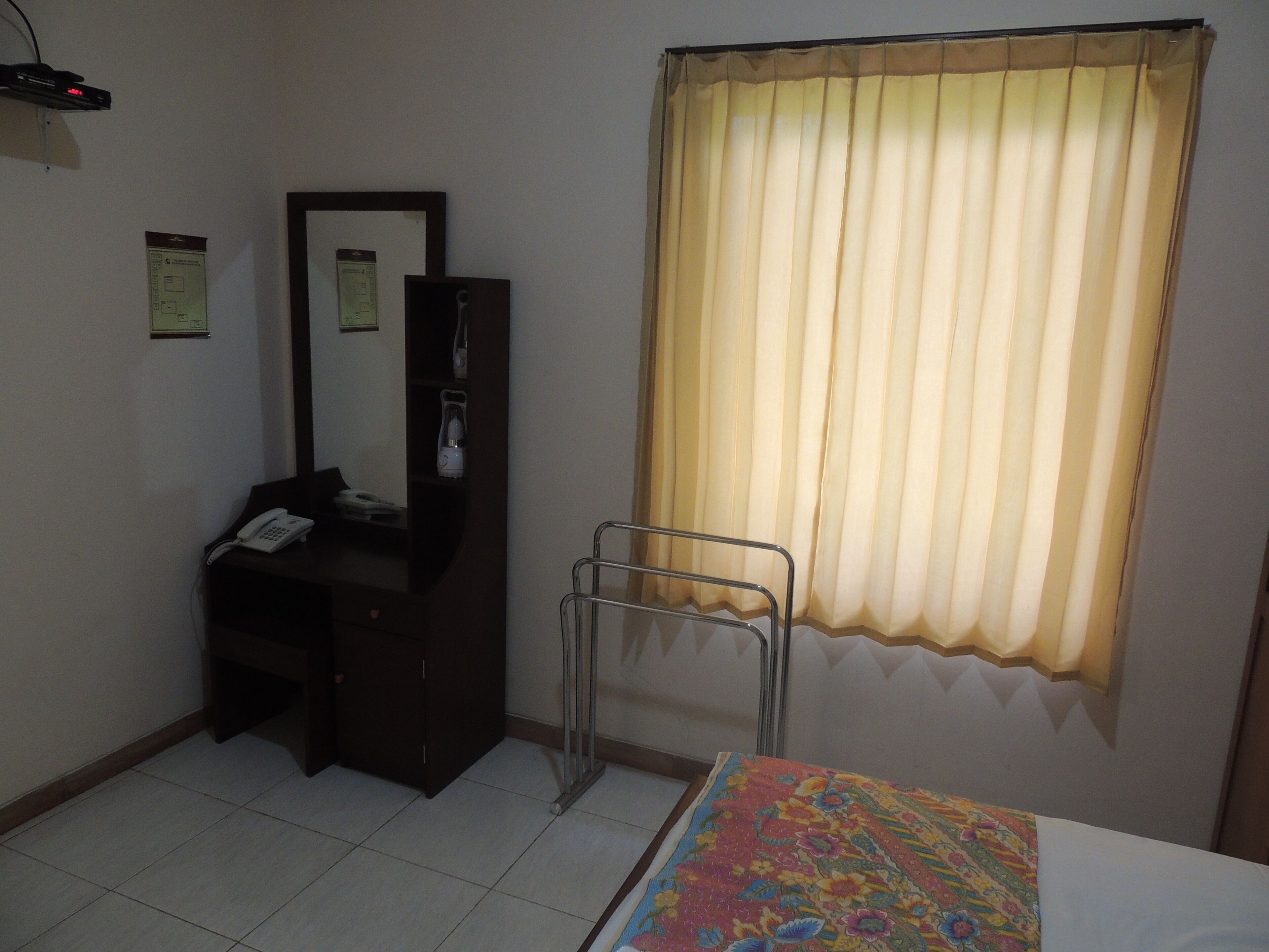 A hotel room with a bed, a desk with a telephone, a mirror, a radiator, and a window with curtains.