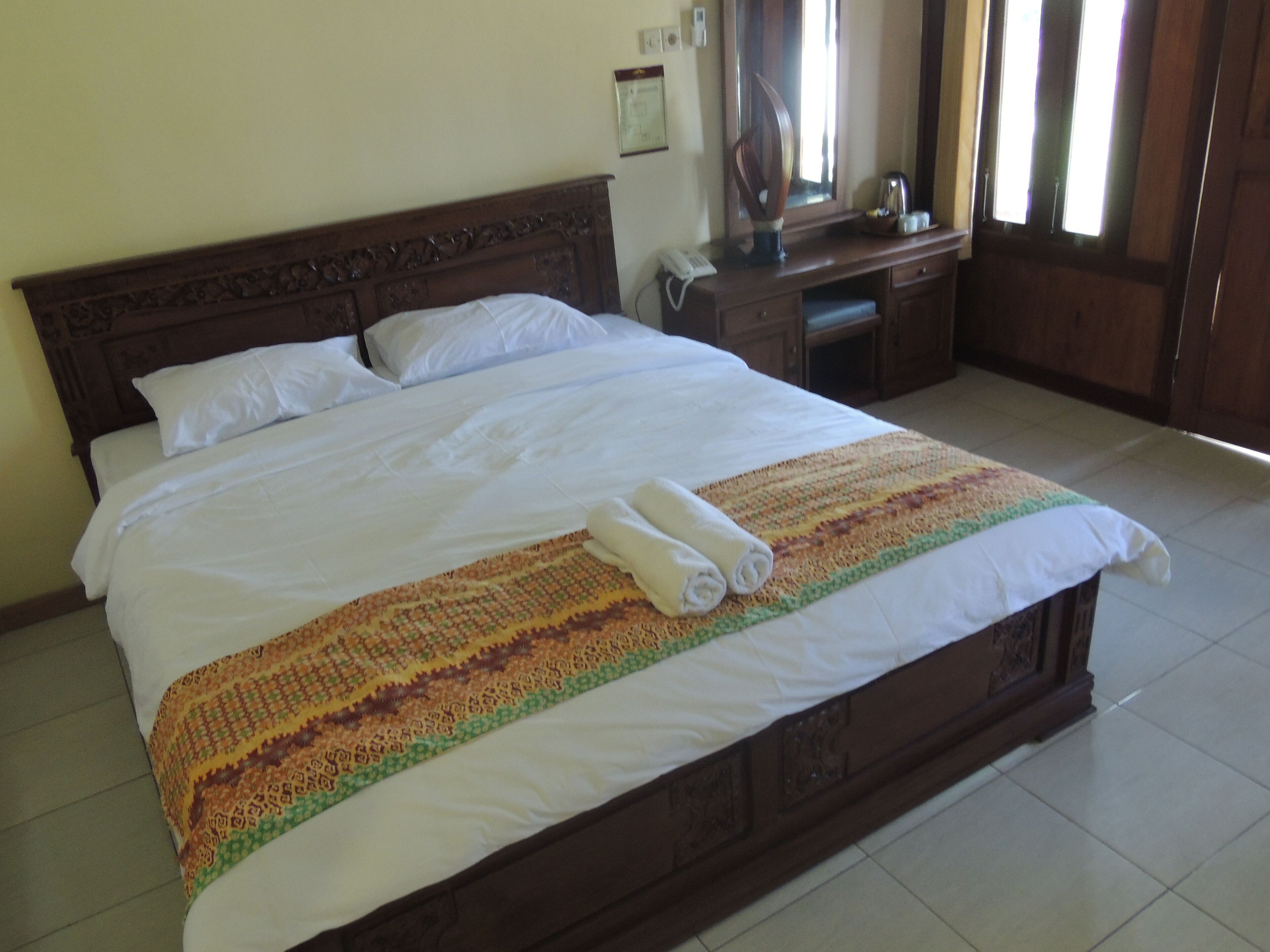 A neatly made bed with a patterned blanket and rolled towels, a wooden headboard, a bedside table with a lamp, and a mirror.