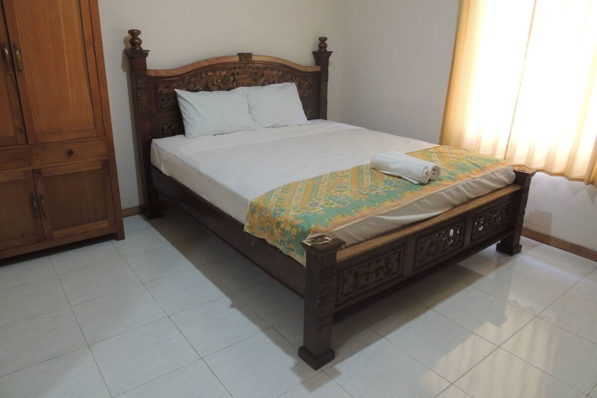 A neatly made bed with a wooden headboard and a patterned bedspread, a wooden wardrobe, and a tiled floor.