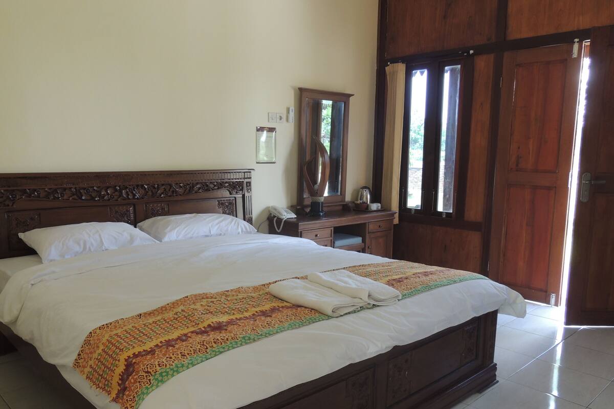 A neatly made bed with a patterned blanket, a wooden headboard, a bedside table with a telephone, and a mirror on the wall.