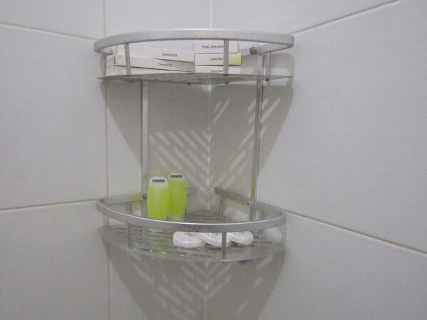 A bathroom corner with a glass shelf holding a dental kit, a bottle of shampoo, and a bar of soap.