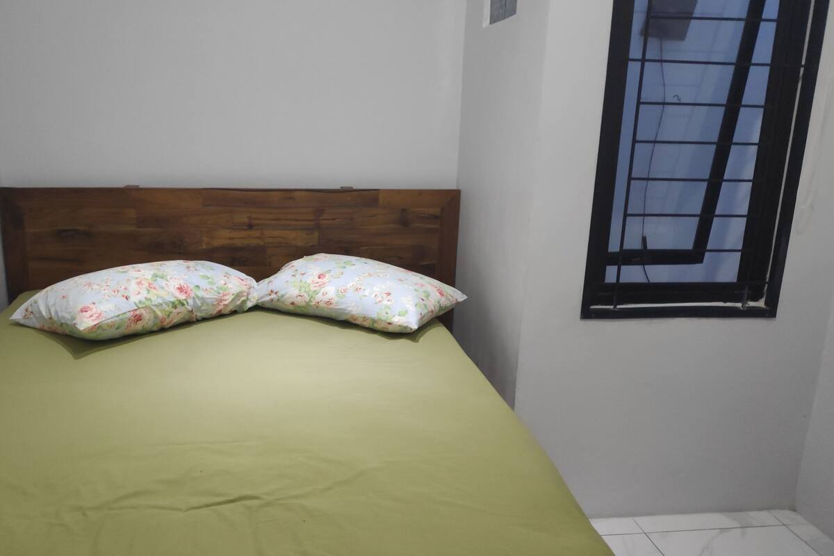 A single bed with a wooden headboard, a green bedspread, and two floral-patterned pillows.