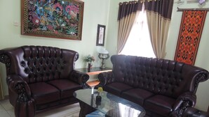 A living room with two dark brown leather sofas, a glass coffee table, a floral rug, a framed painting, and a window with curtains. - Avicenna 2 Guesthouse (Demangsari)