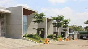 Un edificio moderno con fachada de baldosas, un acceso cubierto y un estacionamiento con algunos árboles y contenedores naranjas.