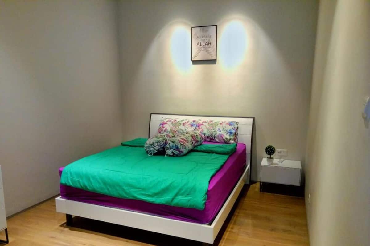 A bedroom with a bed, green and purple bedding, a floral pillow, a small bedside table, and a wall-mounted light fixture.