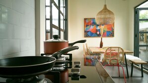 Uma cozinha com fogão, uma frigideira e uma panela sobre o fogão. Há uma mesa de jantar com cadeiras e um quadro colorido na parede.