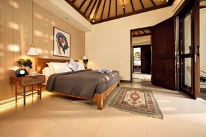 A bedroom with a bed, bedside tables, a lamp, a painting, and a view into another room. - Villa Brabhan Seminyak (Seminyak)