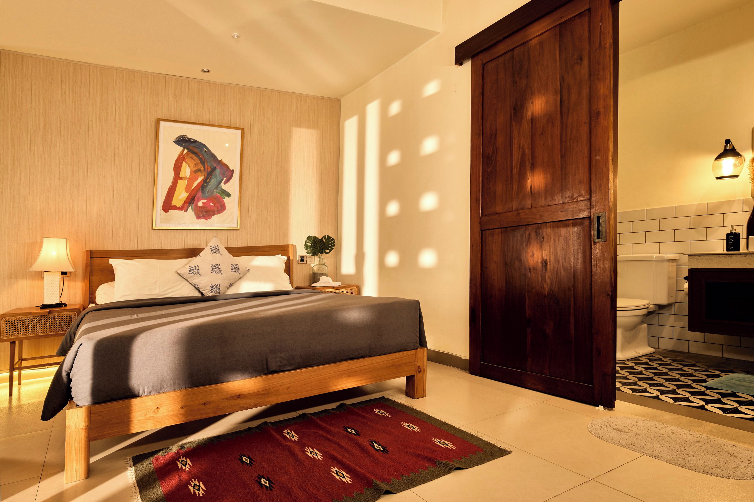 A hotel room with a bed, a nightstand, a lamp, a painting, a large wooden door, and a bathroom visible through an open door.