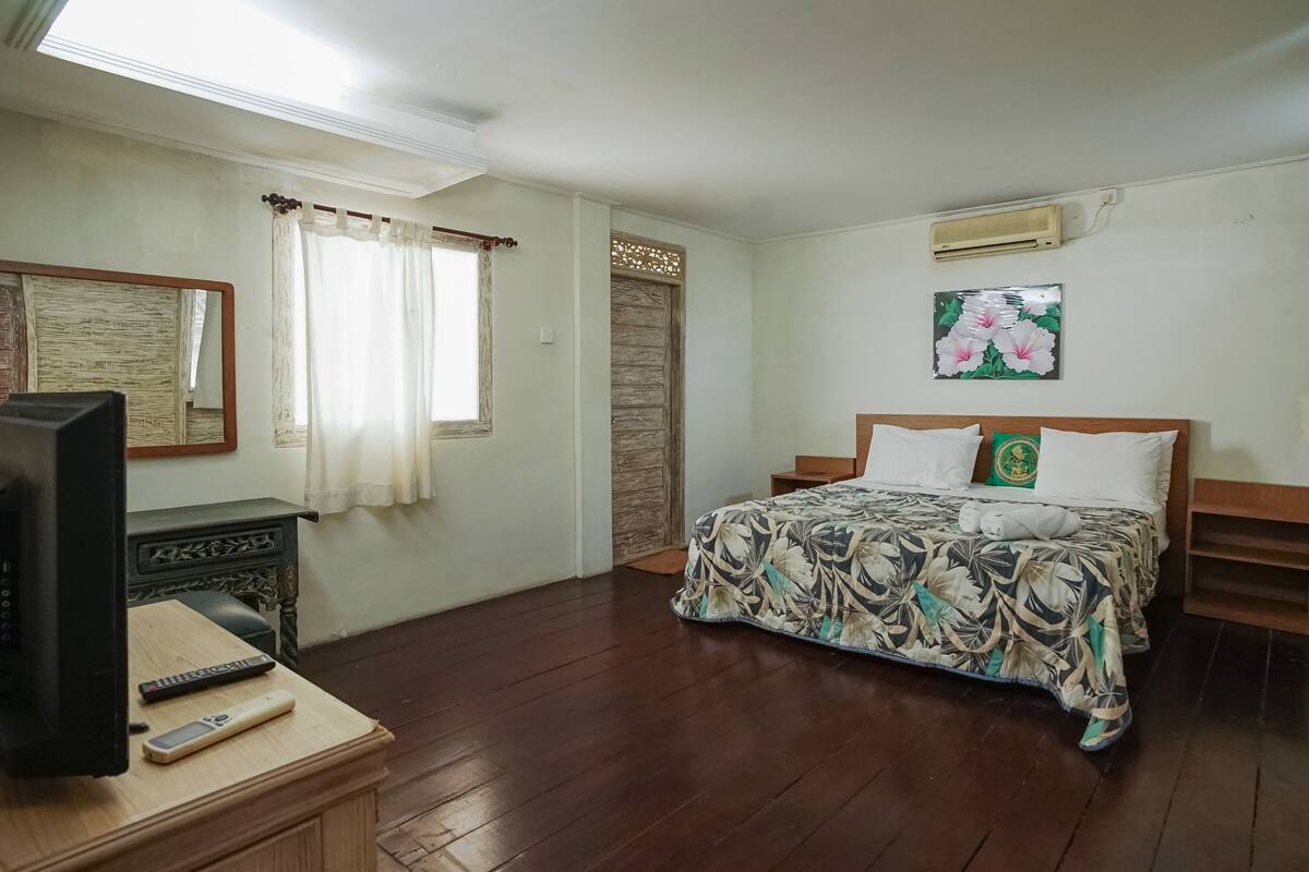 A bedroom with a bed, a television on a wooden dresser, a window with curtains, and an air conditioning unit on the wall.