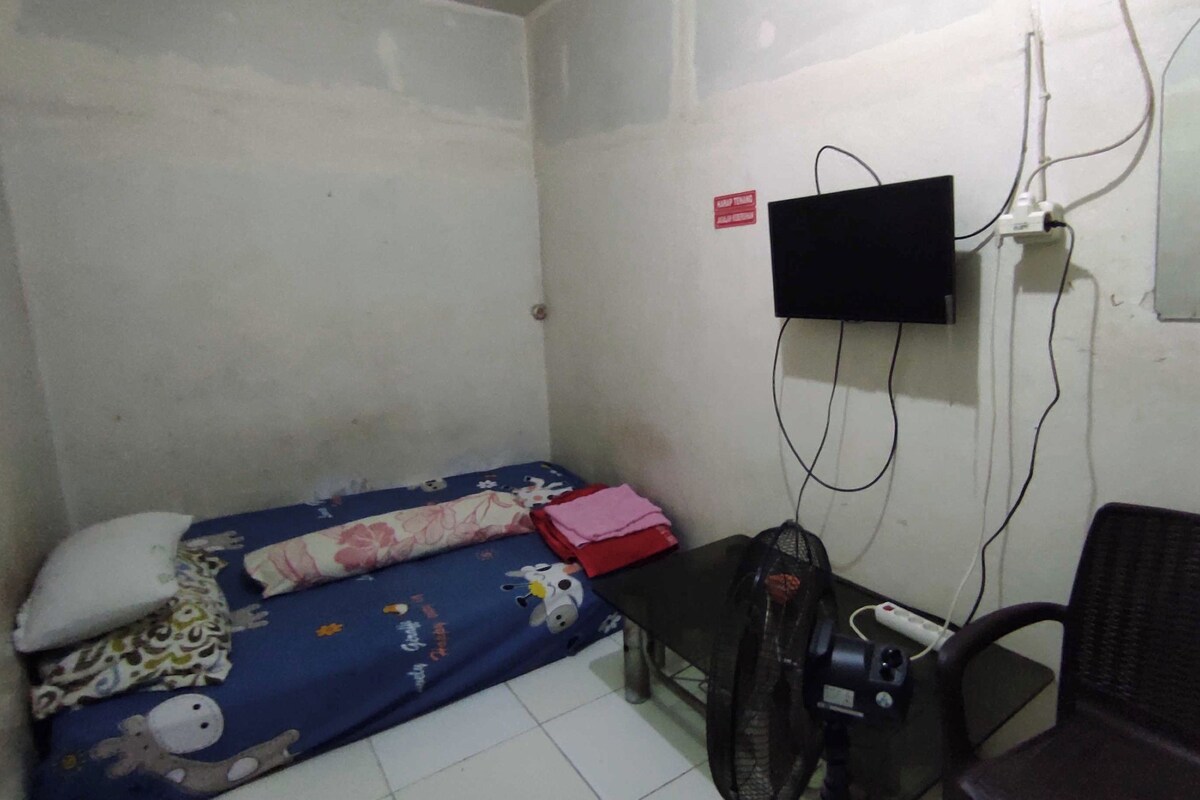 Una stanza piccola e arredata in modo essenziale, con un letto, una sedia, una scrivania e un televisore montato a parete.