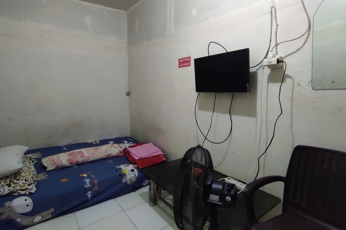 A small, sparsely furnished room with a single bed, a desk, a chair, and a wall-mounted TV.